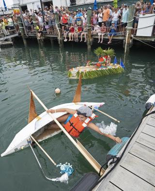 Man in a life vest in a sinking makeshift kayak, crowd watching from a dock.
